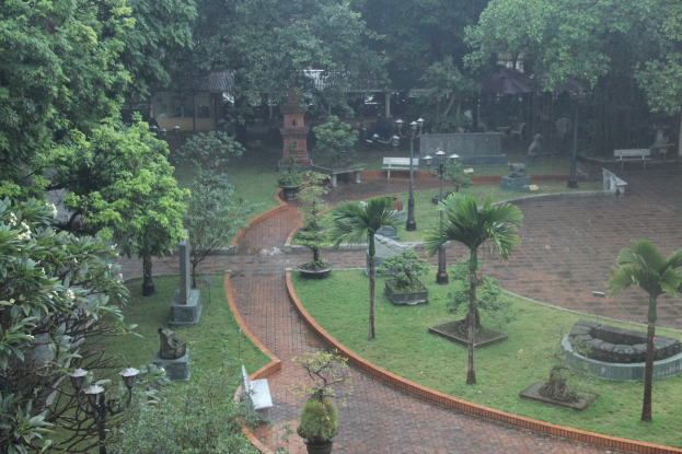 Ein grüner Park mit Bäumen, Pflanzen, Gras, Laternenmasten, Bänken und Skulpturen sowie einem Gebäude im Hintergrund.