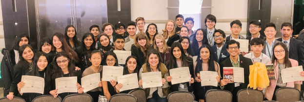 Gruppe von Menschen mit Urkunden bei einer Abschlussfeier, die auf Stühlen sitzen und von einer Wand umgeben sind.