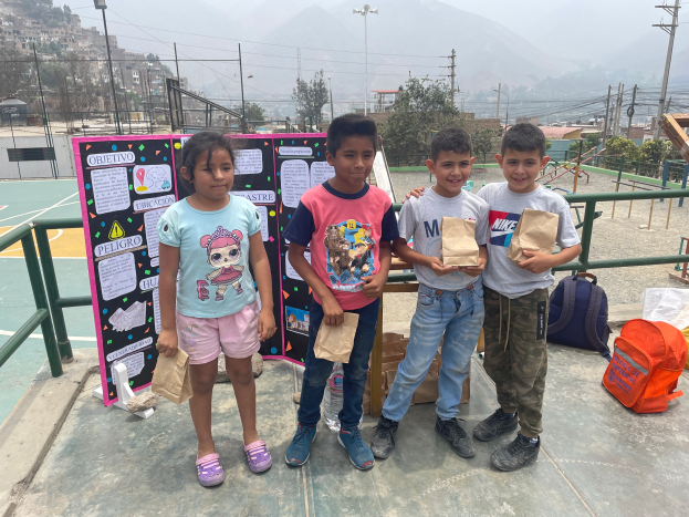 Eine Gruppe von Kindern steht auf einem Basketballfeld, jedes hält einen Papiertüte in der Hand, mit Tüten und Gegenständen rechts und einer Texttafel links, mit Zäunen, Pfosten, Straßenlaternen, Bäumen, Gebäuden, Hügeln und Himmel im Hintergrund.