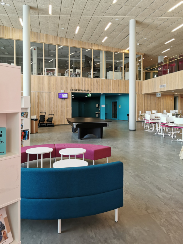 Großer Raum mit Tischen, Stühlen, Sofas, Hockern, Bücherregalen, Säulen, Deckenleuchten und einem wandmontierten Fernseher, der an ein Schulbibliotheksinnere erinnert.