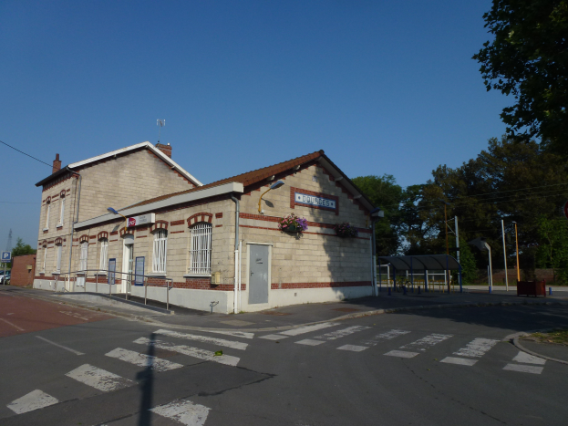 Ein Bahnhofsübergang mit einem Kreuzung vor ihm, umgeben von Gebäuden, Straßenmasten, Straßenlaternen, Strommasten, Stromkabeln, Kraftfahrzeugen, Bäumen und einem klaren blauen Himmel.