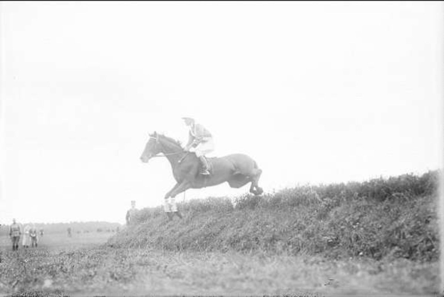 Schwarzes und weißes Foto eines Pferdes und Reiters, die über einen grasbewachsenen Hügel springen, mit dem Pferd in vollem Galopp und wehender Mähne, dem Reiter in einem Helm und einem langen Mantel sowie Zuschauern im Hintergrund.