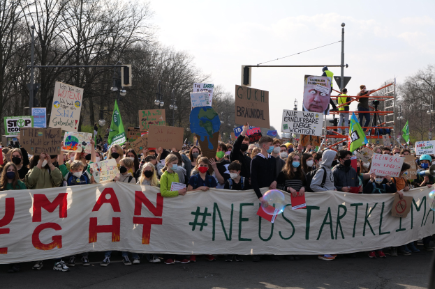 Eine große Gruppe von Menschen marschiert auf einer Straße, hält eine 'Menschenrechte'-Schleife und verschiedene Plakate, wobei einige Masken tragen, unter Bäumen und einem klaren blauen Himmel.