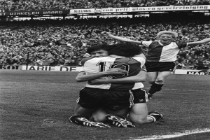 Drei Fußballspieler umarmen sich auf dem Boden und feiern ein Tor vor einer jubelnden Menge in einer schwarz-weißen Stadionszene.