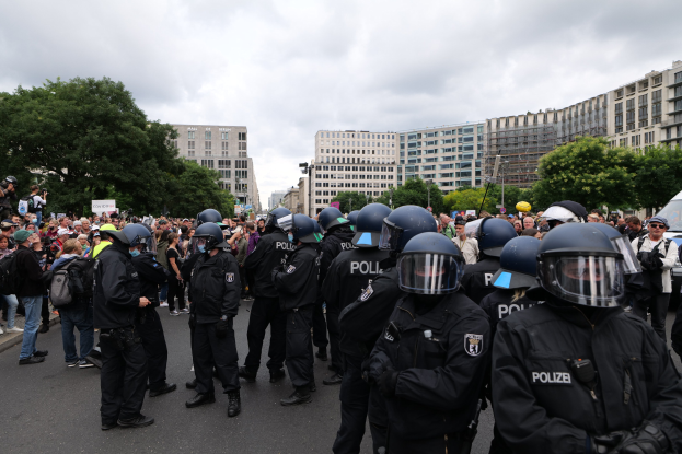 Große Gruppe von Polizeibeamten vor einer Menschenmenge auf einer Straße stehend, umgeben von Bäumen und Gebäuden mit Fenstern, unter einem bewölkten Himmel, während einer Demonstration in Berlin, Deutschland.