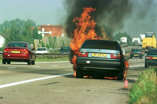 Ein Auto in Flammen am Straßenrand, mit anderen Fahrzeugen in der Nähe, Bäumen, Gebäuden und einem klaren blauen Himmel im Hintergrund und einem Feuerlöscher rechts.