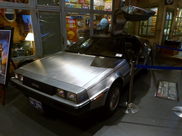 Ein DeLorean DMC-12 aus der Zurück in die Zukunft-Reihe in einem Museum ausgestellt, dessen Türen offen sind und umgeben von Informationsanzeigen, Bändern und diversen Gegenständen.