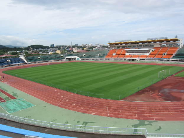 Großes Stadion mit einem Fußballfeld, umgeben von Gebäuden, Bäumen, Hügeln und einem klaren blauen Himmel, mit ein paar Menschen auf dem saftig grünen Rasen.