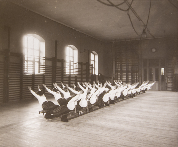 Gruppe von Menschen, die Pilates in einem Fitnessstudio mit einer Uhr an der Wand und Fenstern im Hintergrund machen.