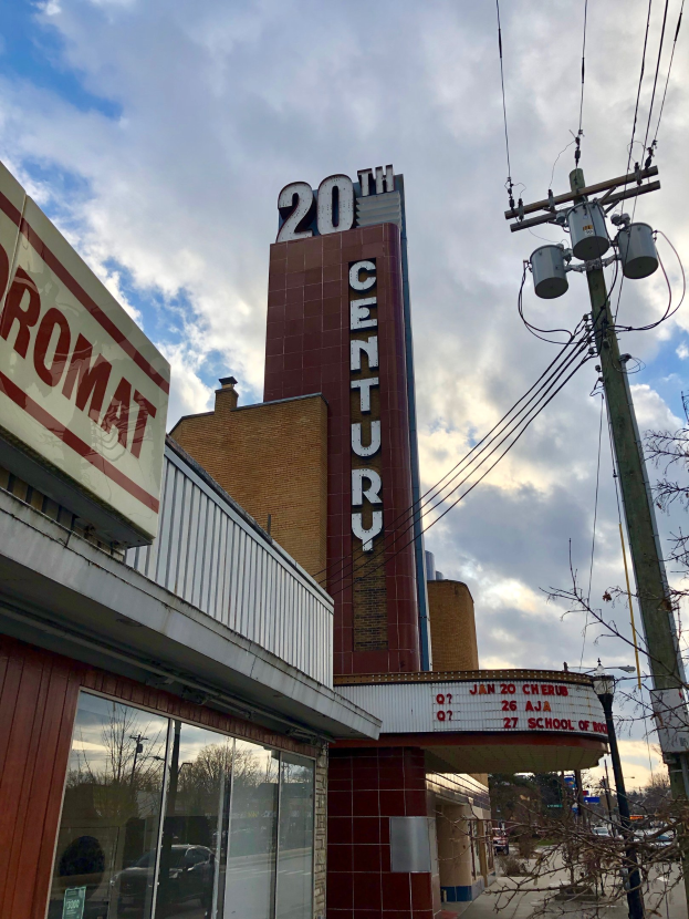 Außensicht des 20th Century Theatre in Detroit, Michigan, mit Glaswänden und Beschriftung, umgeben von Bäumen, Strommästen und Drähten im Vordergrund und einem bewölkten Himmel im Hintergrund.