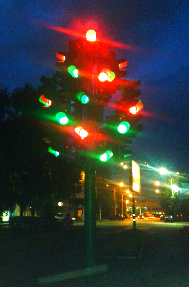 Eine nächtliche Ampel mit leuchtenden grünen und roten Lichtern, umgeben von Fahrzeugen, Bäumen, Gebäuden und Lichtern im Hintergrund unter dem Nachthimmel.