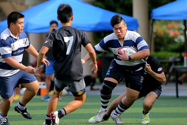 Vier Personen gehen, wobei ein Mann einen Ball hält und blaue Zelte in der Ferne sichtbar sind.