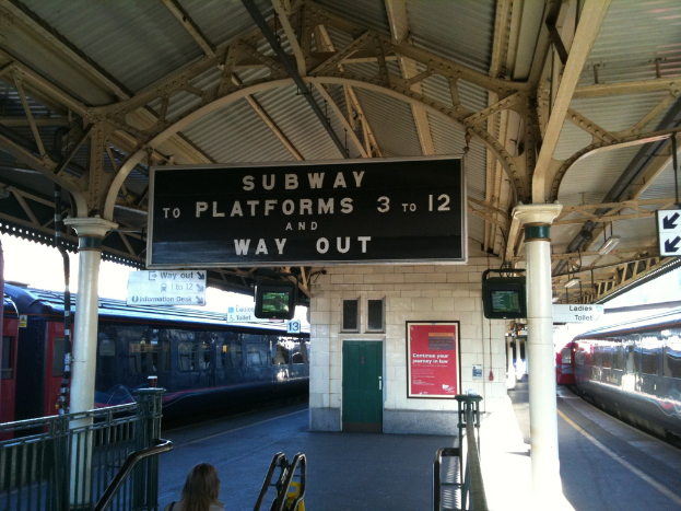 Zwei Züge stehen neben einem Bahnsteig mit zwei Bildschirmen und mehreren Schildern, die von der Decke hängen, eine Person ist in der Nähe der Treppe und eine Tür ist an der Wand zu sehen.
