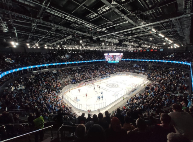Eishockeyspiel in einer großen Arena mit Zuschauern auf den Tribünen und einigen Menschen am Boden, Geländer um die Eisfläche, Überkopfbildschirm und helle Beleuchtung; die Location ist die O2 Arena in London.