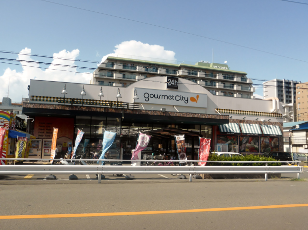 Eine belebte Stadtstraße mit Gebäuden, Straßenmöblierung, Fahrzeugen, Grünanlagen und einem Restaurant, das Speisen in seinem Schaufenster präsentiert.
