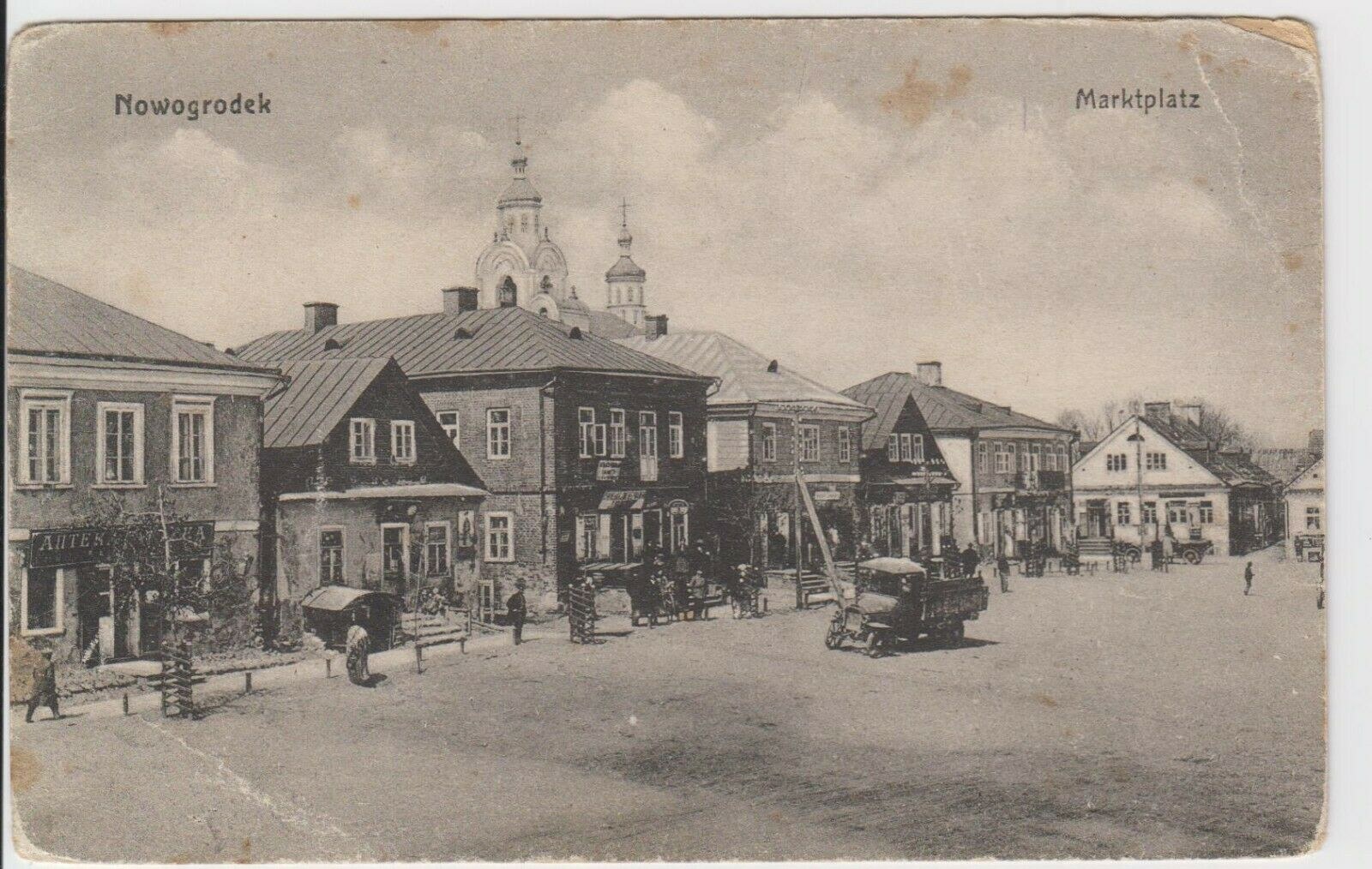 Schwarze-Weiß-Fotografie einer Straße in Nowogrodziec, Deutschland, mit Gebäuden, Menschen, Wagen, Pfölen, Bäumen und Himmel mit Text "Marktplatz in Nowogrodeek, Deutschland" oben und unten.