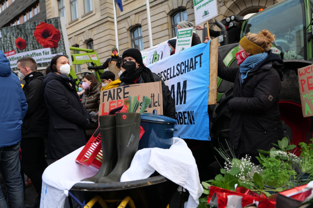 Maskierte Demonstranten mit Protestschildern vor einem Lastwagen mit einem Tisch, Pflanzen, Gebäuden und einer Flagge im Hintergrund.