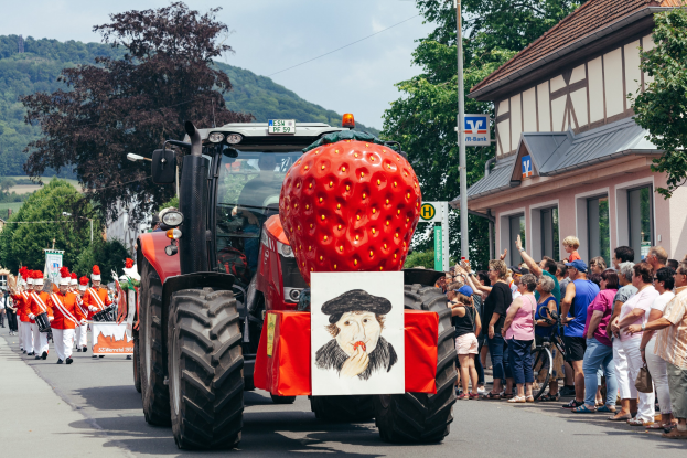Ein großer Erdbeerförmiger Traktor fährt auf einer Straße an einer Gruppe von Menschen auf dem Gehweg vorbei, mit Gebäuden, Bäumen und einem klaren blauen Himmel im Hintergrund.