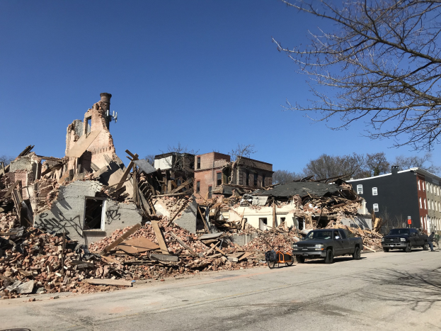 Ein eingestürztes Gebäude an einer Straßenecke mit Fahrzeugen, Passanten, Bäumen und einem klaren Himmel, umgeben von Schutt und Trümmern.