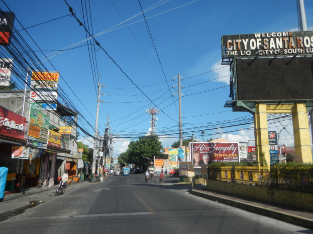Eine belebte Straßenkreuzung mit Fahrzeugen, Radfahrern, Gebäuden, Werbetafeln, Strommasten, Bäumen, einem fernen Turm und einem bewölkten Himmel, mit einer 'Willkommen in Santa Rosa'-Tafel auf der rechten Seite.