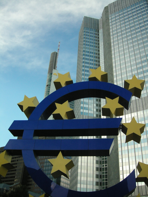 Eurozeichen vor dem Hauptquartier der Europäischen Zentralbank in Frankfurt, Deutschland, mit dem Gebäude umgeben von Bäumen unter einem bewölkten Himmel.
