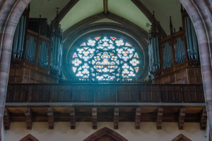 Innenansicht einer Kirche mit einem zentralen Buntglasfenster, das von Holzgeländern und Säulen umgeben ist, sowie verstreuten Gegenständen und einer Wand unten.