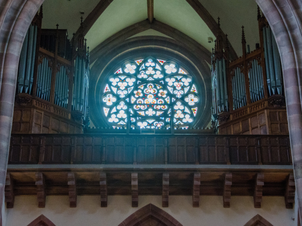 Innenansicht einer Kirche mit einem zentralen Buntglasfenster, das von Holzgeländern und Säulen umgeben ist, sowie verstreuten Gegenständen und einer Wand unten.