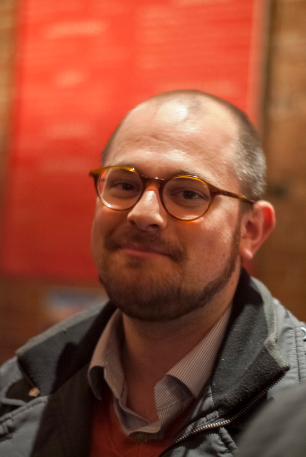 Eine Person in weißem Hemd, schwarzer Jacke und Brille lächelt vor einem braunen und orangefarbenen Hintergrund.