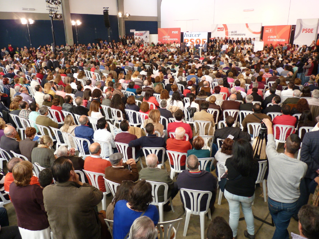 Ein volles Stadion mit vielen sitzenden und stehenden Menschen, eine Bühne mit zwei Rednern auf einem Podium und zahlreichen Bannern und Lichtern in der Nähe.