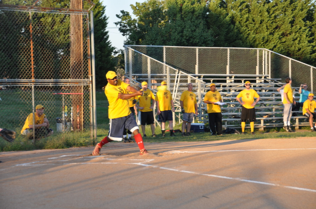 Eine Gruppe von Spielern in gelben Hemden und schwarzen Shorts steht auf einem Feld, wobei ein Spieler im Vordergrund aktiv beteiligt ist, ein Grillzaun und Bäume im Hintergrund.