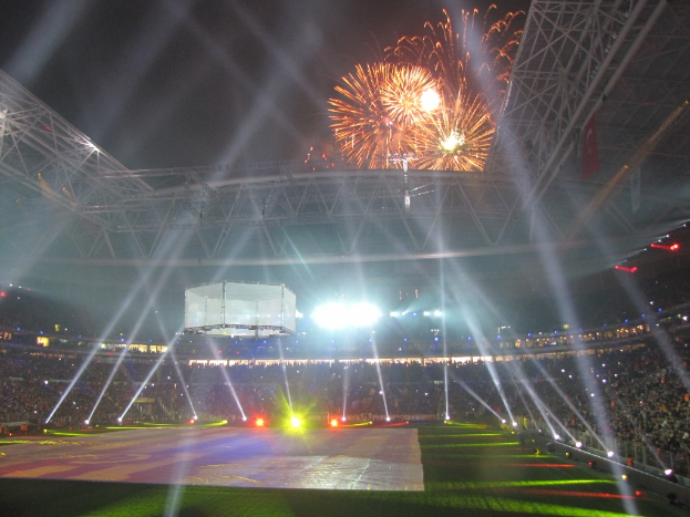 Das Olympische Stadion in London, England, bei Nacht beleuchtet mit einer großen Menschenmenge drumherum, grünem Rasen auf dem Boden und einem Feuerwerk-himmel.