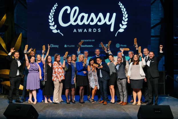 Eine Gruppe von Menschen auf einer Bühne, die Auszeichnungen halten, mit Lautsprechern darunter und einem Hintergrundbild mit der Aufschrift "Classy Awards 2019."