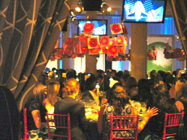 Eine Gruppe von Menschen sitzt an Tischen mit Essen, Fernsehern, Lampen, hängenden Papieren und Vorhängen in einer Art Party-Atmosphäre.