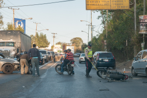 Eine Gruppe von Menschen steht um ein verunglücktes Motorrad auf der Seite einer Straße mit mehreren Fahrzeugen, darunter ein Lastwagen, und einer Hintergrund von Bäumen, Polen, Lichtern und Brettern unter dem Himmel.