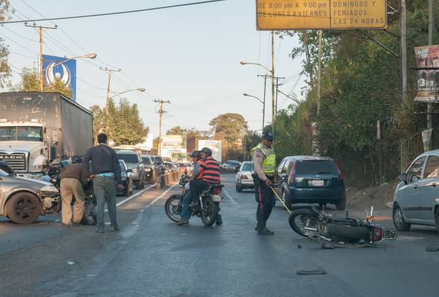 Eine Gruppe von Menschen steht um ein verunglücktes Motorrad auf der Seite einer Straße mit mehreren Fahrzeugen, darunter ein Lastwagen, und einer Hintergrund von Bäumen, Polen, Lichtern und Brettern unter dem Himmel.