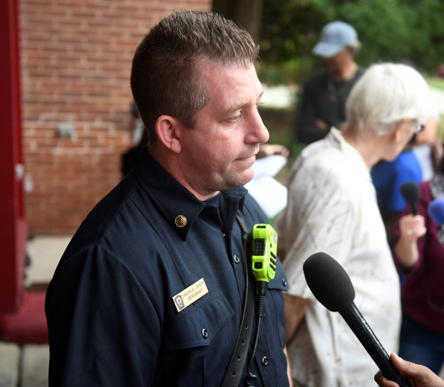 Ein Polizist in Uniform spricht mit Reportern mit Mikrofonen, umgeben von anderen mit Aufnahmegeräten in der Nähe einer Backsteinwand und Bäumen.