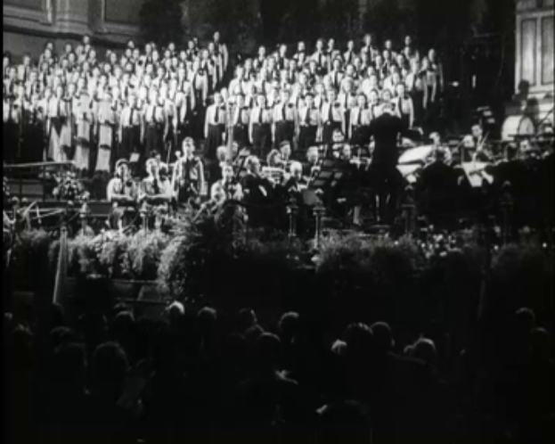 Ein Schwarz-Weiß-Foto einer großen Gruppe von Menschen auf der Bühne, einige halten Musikinstrumente, mit Bäumen und einem Gebäude im Hintergrund und einer Menge unten.
