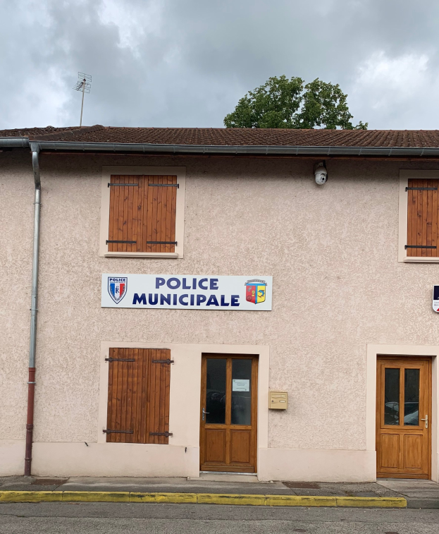Polizeigebäude mit einem Schild, Fenstern, Türen, einer Namenskennzeichnung, einem Rohr, einem Baum und einem bewölkten Himmel im Hintergrund.