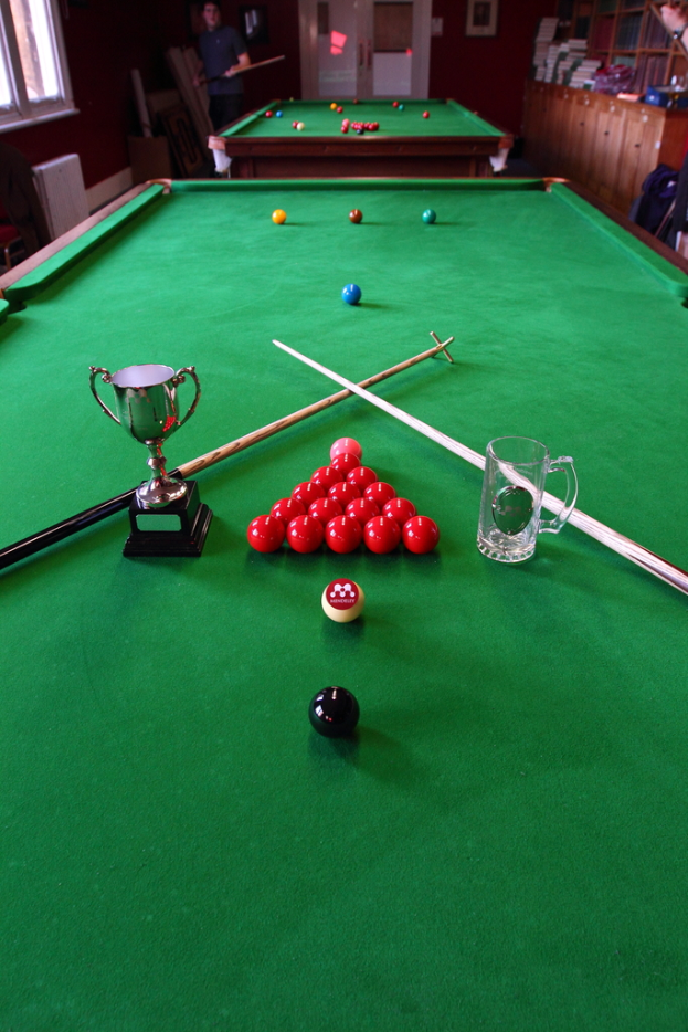 Ein Pokal, ein Glasbecher und mehrere Bälle sind auf einem Billardtisch angeordnet.