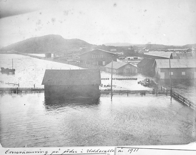 Schwarz-weiß-Foto einer Kleinstadt mit Häusern, Strommasten und teilweise in Überschwemmungswasser versunkenen Booten, umgeben von Hügeln unter einem klaren Himmel, beschriftet mit "1911 Überschwemmungsschäden in St. Petersburg."