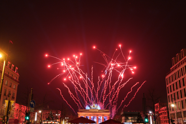 Eine belebte Stadtstraße bei Nacht während des Neujahrsfeierns in Berlin, mit Gebäuden, Bäumen, Laternen, Verkehrszeichen, Zelten, Menschen und einem prächtigen Feuerwerk, das den Himmel erleuchtet.