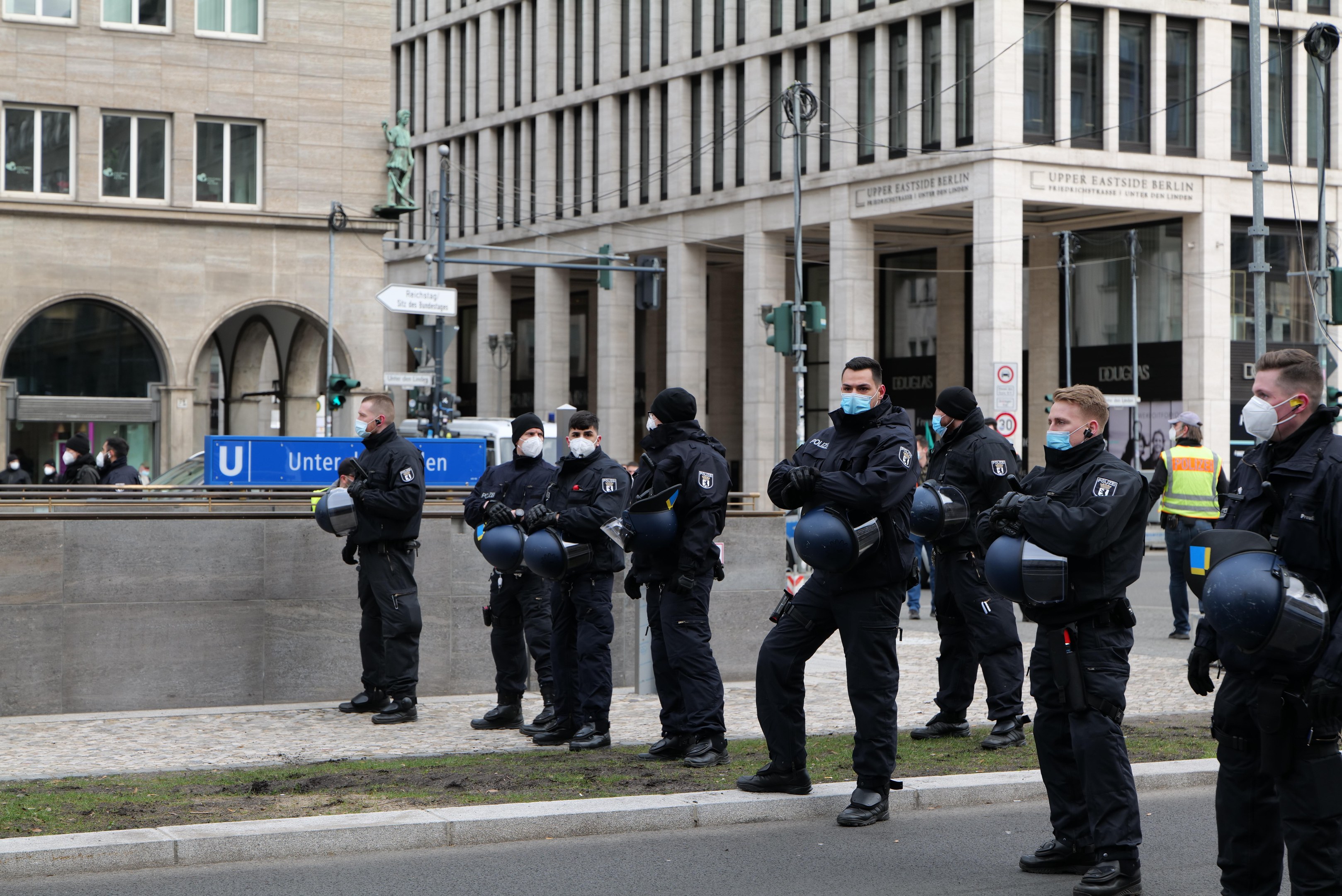 Polizeibeamte in schwarzen Uniformen und Masken vor einem Berliner Gebäude mit Glasfenstern, Säulen und umliegenden Elementen wie Laternen und Verkehrszeichen.