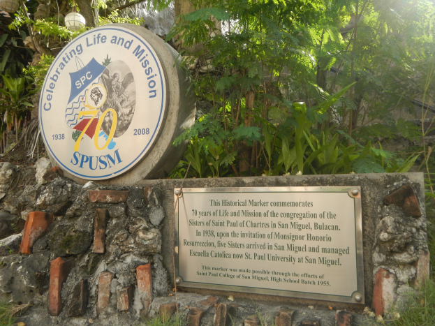 Eine Gedenktafel an einer Steinwand mit der Aufschrift "Leben und Mission feiern" umgeben von Grün und einem blauen Himmel.