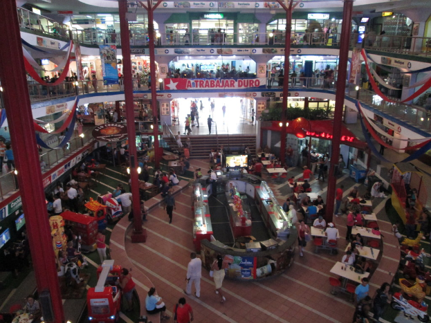 Eine belebte Einkaufspassage mit Säulen, Menschen, Tischen, Fliesenboden, Flaggen, Geschäften, einem Banner, Treppen und einem Namensschild.