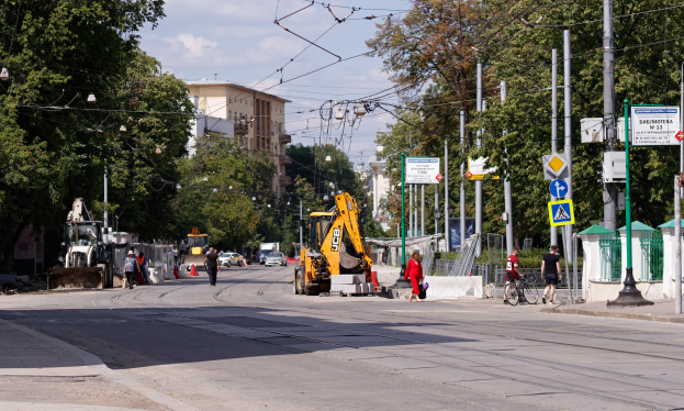 Eine Stadtstraße mit einer Baustelle in der Mitte, mit Fahrzeugen, Fußgängern, einem Radfahrer, Verkehrskegeln, Polen, Schildern, Strommasten mit Drähten, Bäumen, Gebäuden mit Fenstern und einem bewölkten Himmel im Hintergrund.