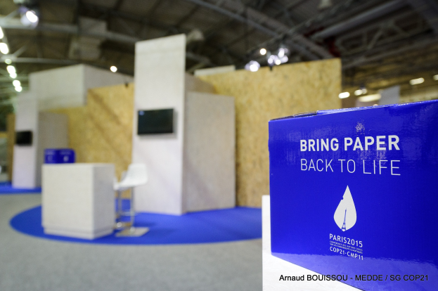 Messestände mit blauer und weißer 'Bring Paper Back to Life'-Schrift auf der rechten Seite, mit unscharfen Stühlen, Tischen, einem wandmontierten Fernseher und Deckenlampen im Hintergrund.