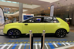 Ein gelbes wasserstoffbetriebenes Elektroauto steht vor einem Gebäude, umgeben von Absperrpollern mit Seilen, Bannern und Schildern, mit einer Person im Auto und einer Gruppe von Menschen in der Nähe, einer Säule, einem Topf mit Pflanze, Deckenleuchten und einer Decke mit Deckenventilatoren.