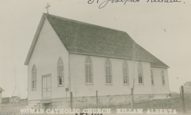 Ein Schwarz-Weiß-Foto von der lutherischen Kirche St. Joseph in Killam, Alberta, das ein Haus mit einem Dach, Fenstern, Türen und einem Kreuz auf dem Dach zeigt, umgeben von einem Zaun, Gras und anderen Häusern im Hintergrund, mit Text oben und unten auf dem Bild.