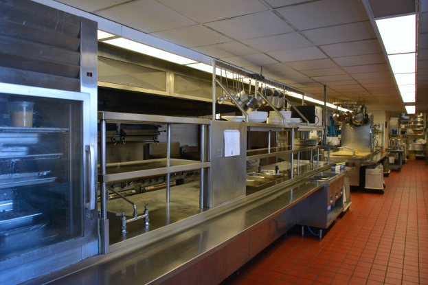 Gewerbeküche mit Edelstahl-Theke, elektrischer Deckenbeleuchtung, hängenden Utensilien, einem Spülbecken, Wasserhähnen, Schränken und diversen auf Gestellen angeordneten Gegenständen.