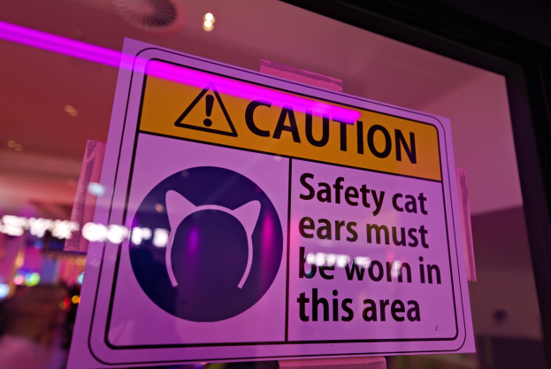 Achtungsschild in einem Ladenschaufenster mit der Aufschrift "Achtung: Sicherheitskatzenohren müssen in diesem Bereich getragen werden" auf einem farbigen Hintergrund.